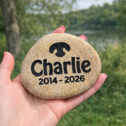Hand holding a engraved stone with 'Charlie' and dates 2014-2026, outdoors.